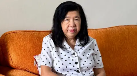 Susanna Liew, wearing a white top with black polka dots, is seated on a brown sofa and looking straight at the camera