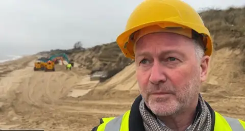 Andrew Turner/BBC A man with a grey beard and a yellow hard hat stands on a beach. In the background is a yellow digger.
