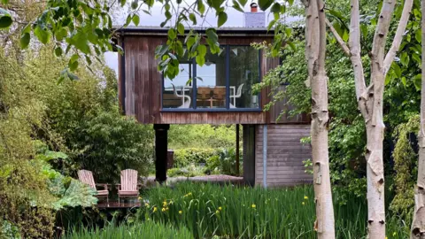 A timber house on stills above a lake. There's greenery all around and the house has huge windows to overlook the views. Inside, a table and chairs can be seen by the window.