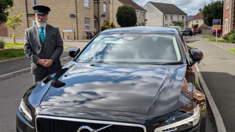 Hillier Funeral Service The front of a black, shiny limo parked in a residential area. To the side, the driver stands next to it smiling at the camera. He is wearing a chauffeur-type outfit, complete with hat and a blue tie.