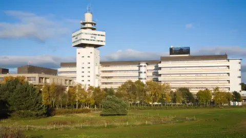 Getty Images A big building spans across the whole image. On the left there is a tower, which has a mast on the top, and a sign which says BT. The building is white, and has lots of smalls windows going across it. 