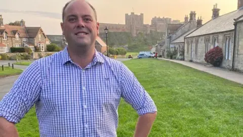 Guy Renner Thompson, a councillor in a blue shirt stands with hands on hips in front of Bamburgh Castle 