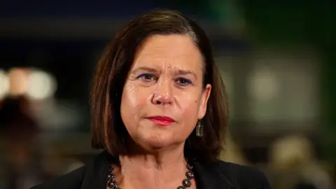 PA Media Mary Lou is looking serious, mid conversation. The background is blurred out and her face is in shot. She has brown eyes and has gold stud earrings on. She has brown shoulder length hair and red lipstick on.