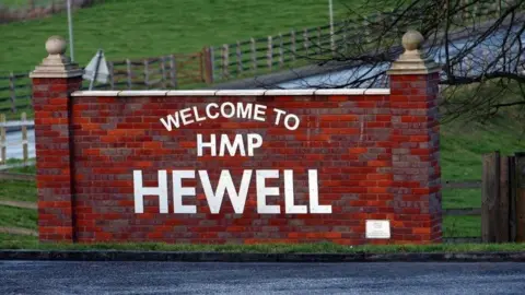 A red brick wall at the side of a road, saying 'Welcome to HMP Hewell' in white lettering. Wooden gates and grass are at the side of the road.
