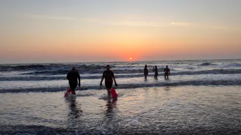 BBC / Amanda White At sunrise, five women emerge from a chilly dip in the North Sea. The sun is rising in the distance