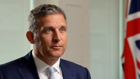 Prisons minister James Timpson is pictured on the left hand side of the frame wearing a navy blue suit jacket, white shirt and pale blue tie. A Union Jack flag can be seen on the right hand side of the frame.