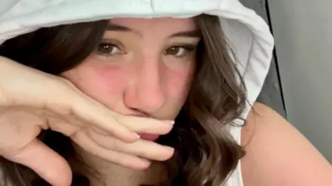 Family photo Levi Davies is a young woman with long dark hair and brown eyes. She is wearing a white hood with her hand over her mouth. 