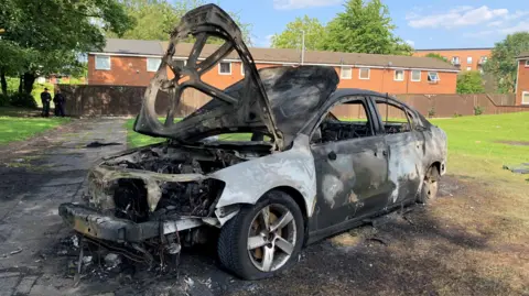 A car which has been burnt out and and is completely destroyed is on the grass on a residential street. 