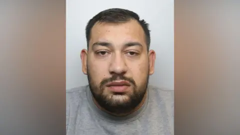 A police mugshot of a man in his twenties, with dark hair and a short beard. He is wearing a grey t-shirt and staring directly into the camera.