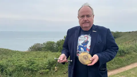Alex Fearn standing on a grassy coastal hillside at Portlet overlooking the sea under an overcast sky. He is positioned on the right side of the frame, facing the camera, wearing a dark blazer over a graphic T-shirt and holding two metal objects - one circular brass disc and one rod-like piece. The ocean and shipwreck site lies behind him.