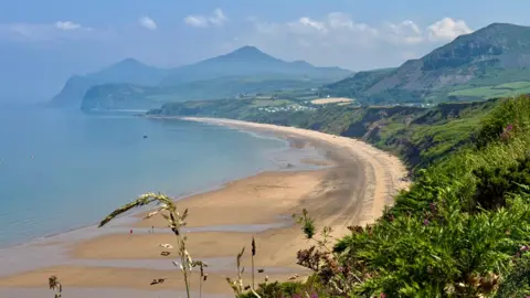 Weather Watchers/SusySunshine Beach in curve with lush green vegetation above and mountains in the background