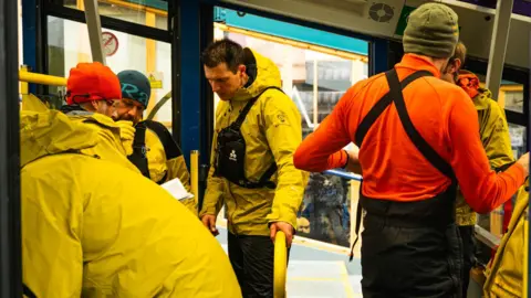 Angus Trinder/Cairngorm Mountain Six CMRT volunteers wearing bright clothing inside a cabin on the Cairngorm funicular.