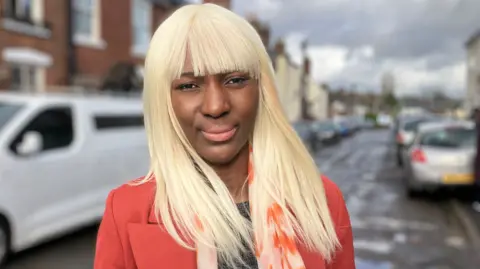 Celia has long platinum blonde hair with a fringe. She is standing in a wet road with houses either side and cars parked on both sides. She wears a black top and orange suit jacket