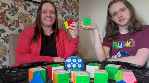 Louise and Thomas are sitting at a table with a range of brightly coloured Rubik's Cubes in front of them. Louise is in her 40s with long brown hair, Thomas is 15 with shoulder length brown hair and wearing a t-shirt. 