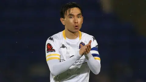 Getty Images Michael Clunan is standing on a football pitch. He has short brown hair and is wearing a white football T-shirt which has branding on it. He is clapping his hands while walking.