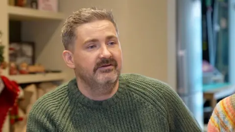 A man in a green sweatshirt looking to the right of the camera. He has a dark brown moustache and grey beard, as well as greying hair. 