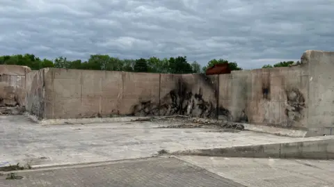SAM READ/BBC The picture shows the burnt out remains of a building where only external walls remain. These concrete structures, with damage and scorch marks can be seen across the middle of the picture. Below is a concrete floor. Above the top of a tree line can be seen and above that a cloudy overcast sky.