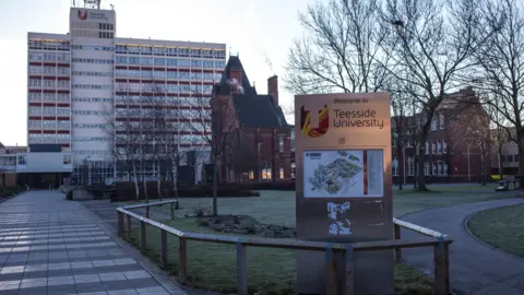 Kampus Getty Images Teesside University. Sebuah tanda berisi peta kampus berdiri di halaman luar salah satu gedung universitas. Ada sejumlah pohon di belakangnya, semuanya tanpa daun.