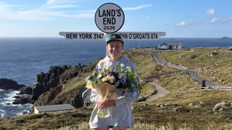 Erin More (@moremomentz) A picture of Ella Fryer. She is holding a bouquet of flowers in front of a white sign at Land's End.