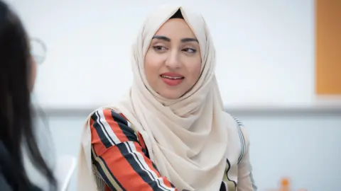 Syeda Batool sitting down, looking at a person to her left. She has a scarf covering her head, and is wearing a striped long sleeved top. 