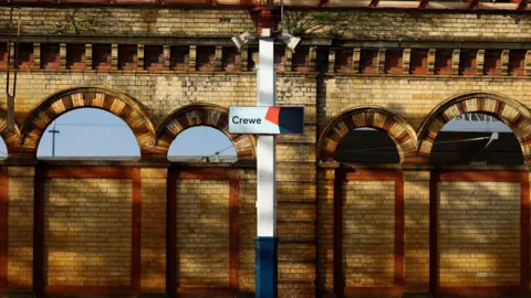 A sign saying Crewe at Crewe Railway Station. It is on a white pillar, with ornate pale-brick station walls behind it. 