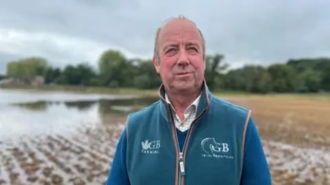 Graham Bowsher, wearing a blue fleece gillet over a jumper and shirt. In the background is a waterlogged muddy field.