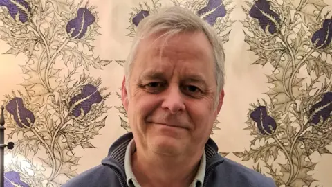Iain Smith Iain Smith, who has grey hair and is wearing a blue top, stands looking into camera in front of thistle wallpaper 