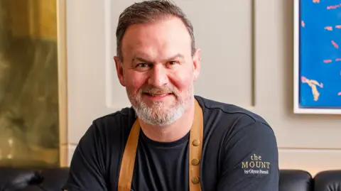 A man with short dark hair, a grey beard, black shirt with "The Mount by Glynn Purnell" on the left sleeve, sits and smiles at the camera.