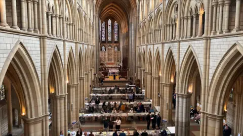 BBC A photo taken of Truro Cathedral with rows of horizontal desks placed along the main section of the cathedral. People are sat at them with sheets of paper in front of them. At the end of the cathedral is a large stained glass window.