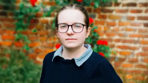 A woman with her hair tied back and wearing glasses, looks into the camera. She is wearing a navy jumper and shirt. A brick wall and bushes can be seen in the blurred background behind her.