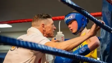 A boxing club coach, on the left, talking to a young boxer, on the right of the image. The coach is wearing a white T-shirt and has a towel around his neck. The fighter is wearing blue boxing gloves and blue protective headgear.