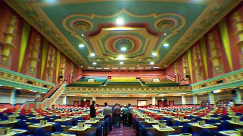 Project Gaumont Inside interior of a huge arts venue. There are tables and chairs dotted on the floor area with blue seats. There are circle, upper seating at the top. It looks like a grand palace with the interior, with colours of red, bright yellow and a blue and yellow ceiling.