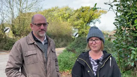 Rick MacMahon, in a brown jacket, grey jumper and shirt and wearing glasses, and Laura Gould, who wears glasses and a grey beanie, stand facing the camera. Behind them is a track and vegetation. 