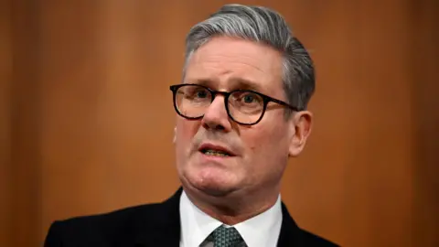 Sir Keir Starmer, dressed in a black suit jacket, white shirt, and green patterned tie, stands against a plain warm brown background. 