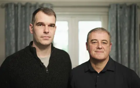 Michael and David are standing together indoors in front of a softly lit window with pale curtains. Michael, on the left, is wearing a dark zip‑up sweater over a grey t-shirt. David, on the right, is dressed in a dark collared shirt.