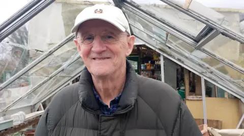 BBC Tom Haire, with white cap and grey jacket, standing inside greenhouse with glass panels.