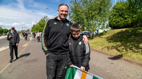 A man and a boy wearing Celtic jackets stand together on a sunny street before a match, with the boy holding an Irish flag as fans walk in the background.