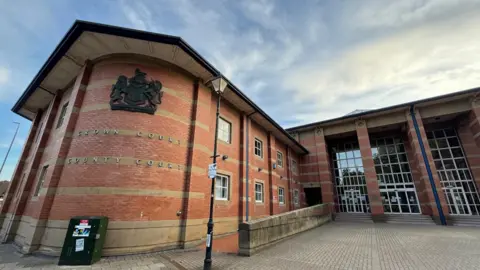 Stafford Crown Court, a brick-built building with large windows stretching from the ground to the roof. There is a crest on a curved section of the building and a ramp leading up to its main entrance.