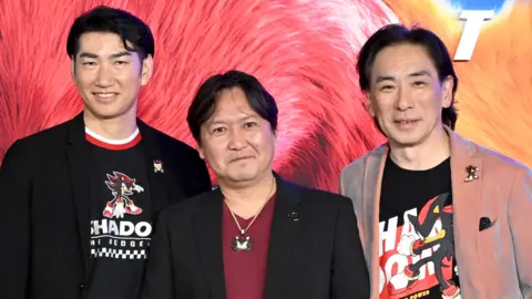 Getty Images Three Japanese men stand together smiling for a photograph at a film premiere. Behind them is a backdrop featuring characters in the Sonic movie. The three men are each wearing an item of clothing featuring Shadow the Hedgehog, who features in the film. Satomi and Utsumi have Shadow on a t-shirt as well as lapel badges, while Iizuka has a Shadow necklace.