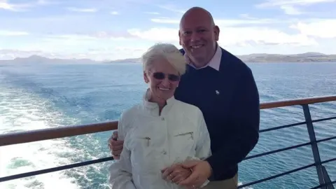 Brian Scott Brian and Aileen Scott are pictured on the deck of a cruise ship with the ocean in the background. They are both staring at the camera and smiling.