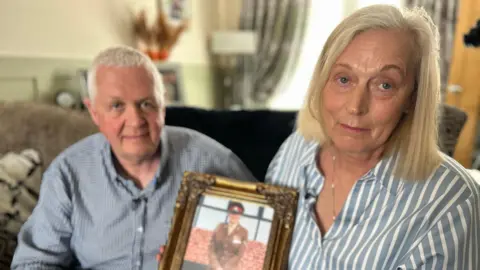 Malcom and Amanda sitting on a living room sofa, holding up a framed picture between them of Nadine wearing a brown military uniform. They are both looking at the camera.