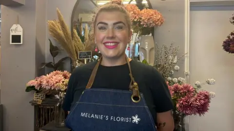 A woman with bright pink lipstick, pink dangly earrings and light pink hair tied back in a bun stands in front of a floral display. She wears a black t-shirt and a denim-style apron with white lettering that reads: Melanie's Florist.