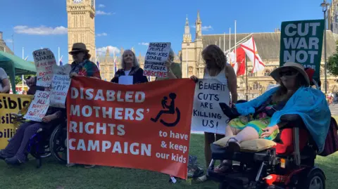 Joy Dove stands behind a large red sign for the Disabled Mothers' Rights Campaign. She is joined by other campaigners on Parliament Square who are protesting the government's welfare reforms.