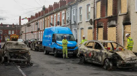 Reuters Three burnt out cars on a street of terraced houses in east Belfast are inspected by gas engineers