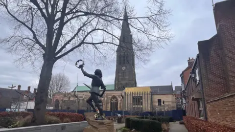 A statue of Richard III raising a crown with Leicester's spired cathedral in the background