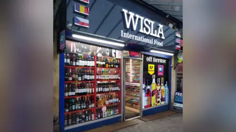 LDRS A brightly lit convenience store called “WISLA International Food,” with an open door, shelves of snacks inside, and a window display filled with bottles of alcohol.