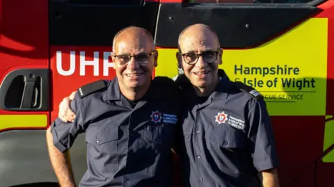 Dave and Paul Minall smiling in front of a Hampshire & Isle of Wight Fire and Rescue Service vehicle. They have their arms around each other's shoulders.