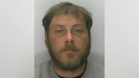 A police mugshot of Ashley Quinton. He has blue, bloodshot eyes, a moustache, a beard and short brown hair.