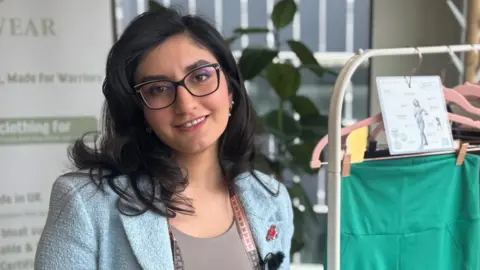 Roya Rasouli is smiling directly at the camera. She is standing next to a collection of leggings designed to help those who struggle with endometriosis. She is wearing a light blue blazer and has a tape measure around her neck. 
