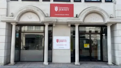 BBC The Government of Jersey building - a stone and glass-fronted edifice, with signs on it 